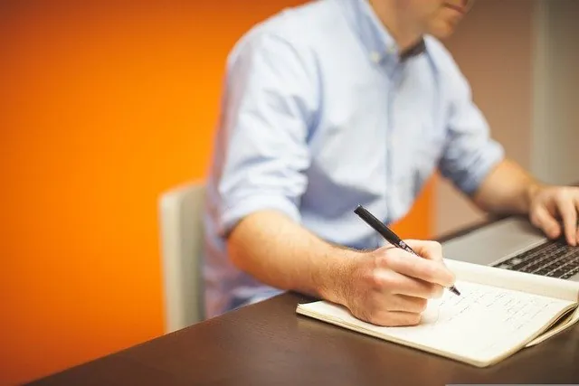 Man in a light blue shirt writing in a notebook beside a laptop against an orange wall, planning an employee retention claim