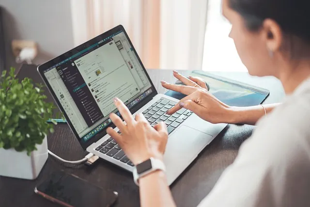 Woman typing on a laptop at a wooden desk with a plant and phone nearby, running a woman-owned business from home