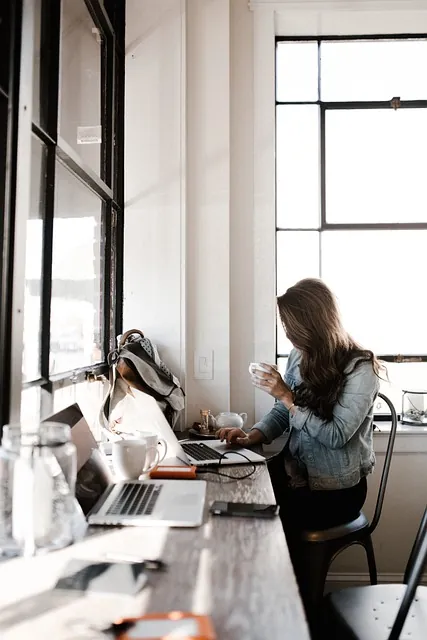 Woman sipping coffee while working on a laptop at a sunlit window counter, the flexibility of women-owned certification